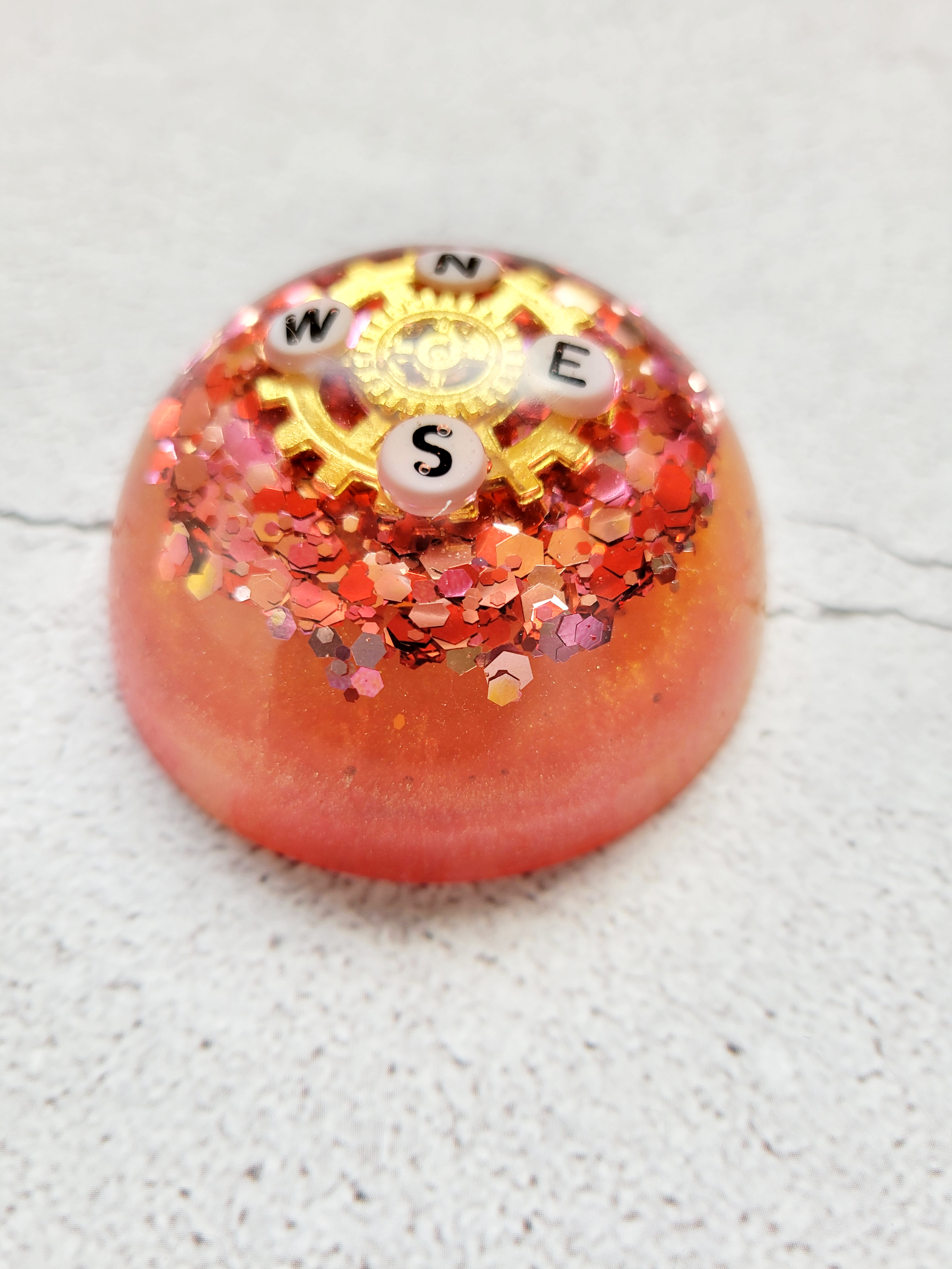 A side view of a reddish orange half bubble with a gold gear with the compass directions N, E, S, W on it, laying on a bed of red and orange glitter.