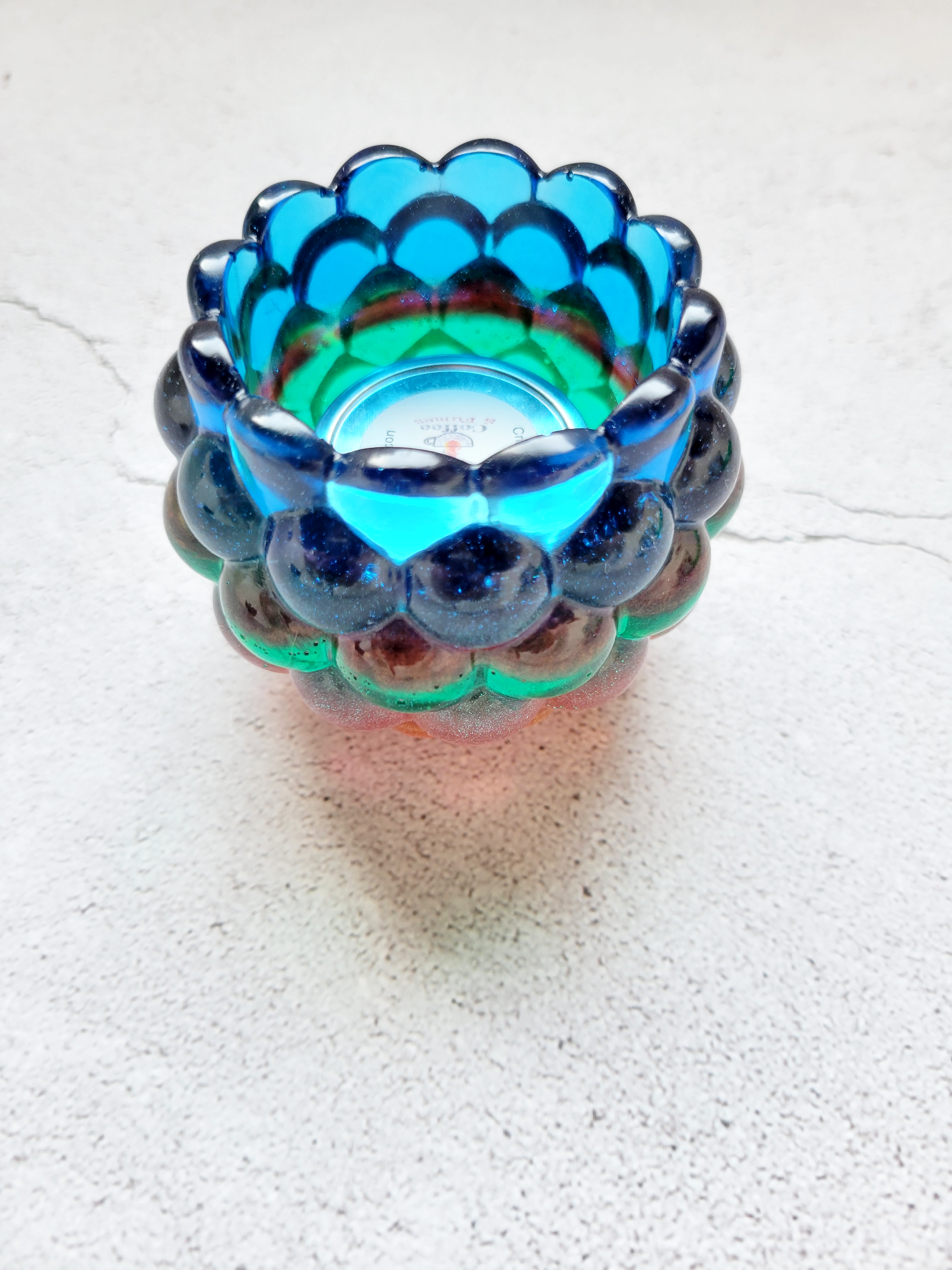 A side view of a tealight candle holder with a bubbled look. It's layered with colors of blue, red, green, orange. There is a tealight candle inside to show size comparison.