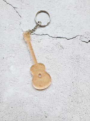 An acoustic guitar keychain. It has a silver chain and ring hoop. It's transparent glittery gold.