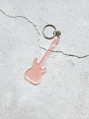 A keychain in the style of an electric guitar. There is a silver chain and hoop connected to the guitar head. It's a pale translucent pink.