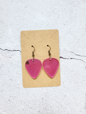 A pair of guitar pick earrings on a cardboard backer card. The earring hooks are antique colored. The guitar picks are translucent dark pink with fine silver glitter throughout. The left pick has a small dark brown spot along the outer left side.