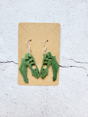 A pair of earrings on a cardboard backer card in the style of t-rex skulls. The hook earrings are silver in color. The skulls are green.