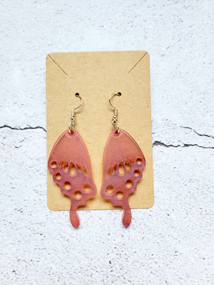 A pair of earrings in the style of butterfly wings on a cardboard backer card. The hook earrings are silver in color. The wings are pink and purple toned.