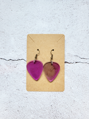 A pair of earrings in the style of guitar picks on a cardboard backer card. The earring hooks are antique colored. The picks are a translucent purple with fine glitter mixed throughout. The right pick has a patch of clear on the left half.