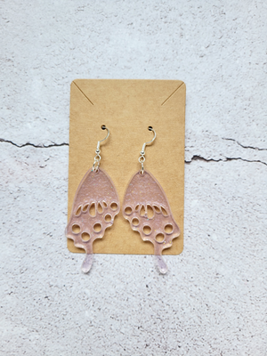 A pair of butterfly wing earrings on a cardboard backer card. The earring hooks are silver in color and the wings are a transparent pink with fine silver glitter mixed through.