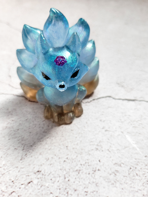 A front view of a kitsune fox figure with nine tails. It is blue and tan, with black painted eyes and nose, with a purple flower on its head.