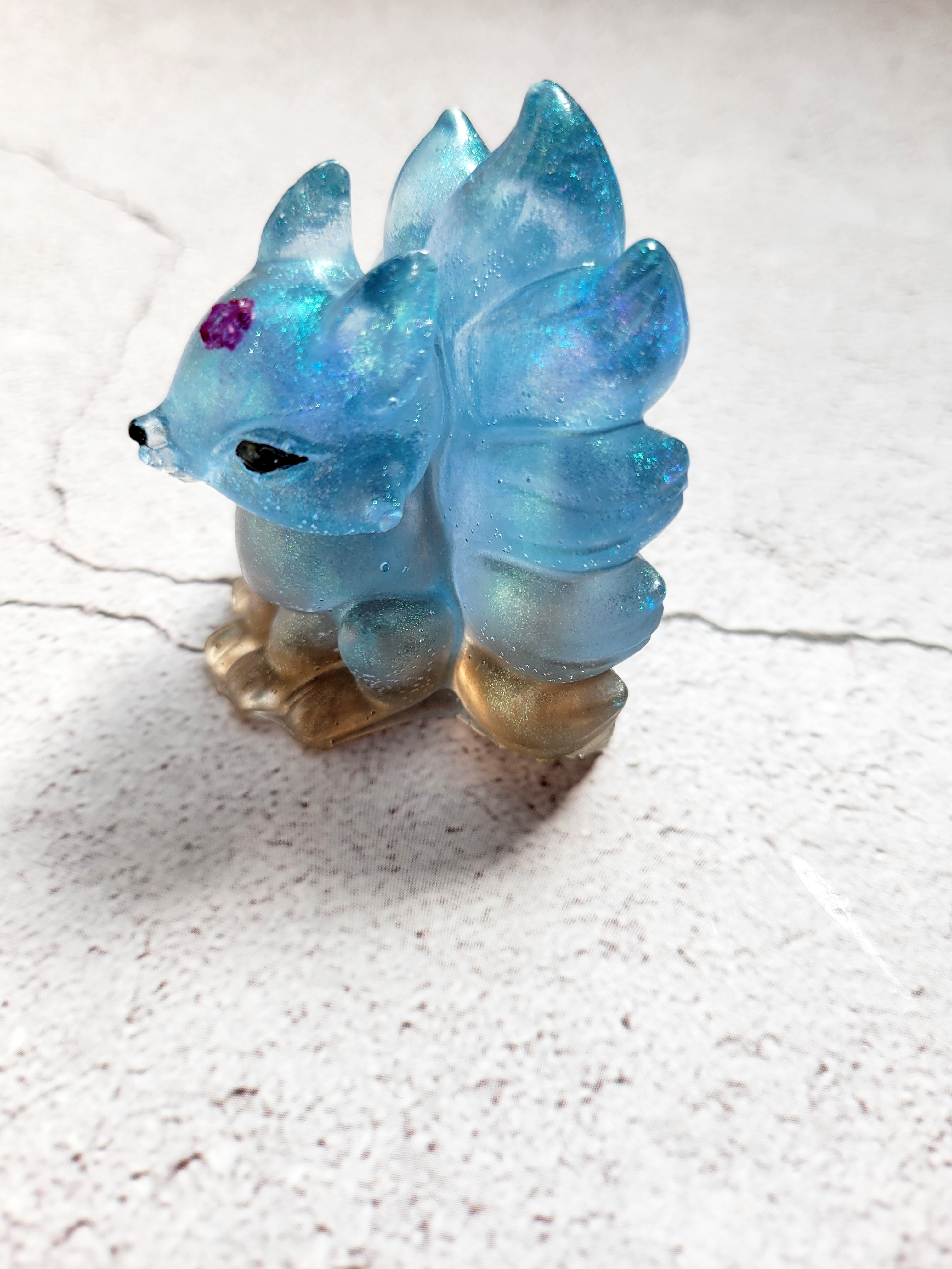 A side view of a kitsune fox figure with nine tails. It is blue and tan, with black painted eyes and nose, with a purple flower on its head.