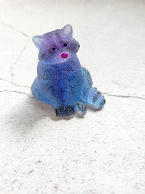 A front view of a sitting raccoon figure. He's a mix of blues and purples with black painted eyes, feet, tail and fur spots. His nose is painted red.