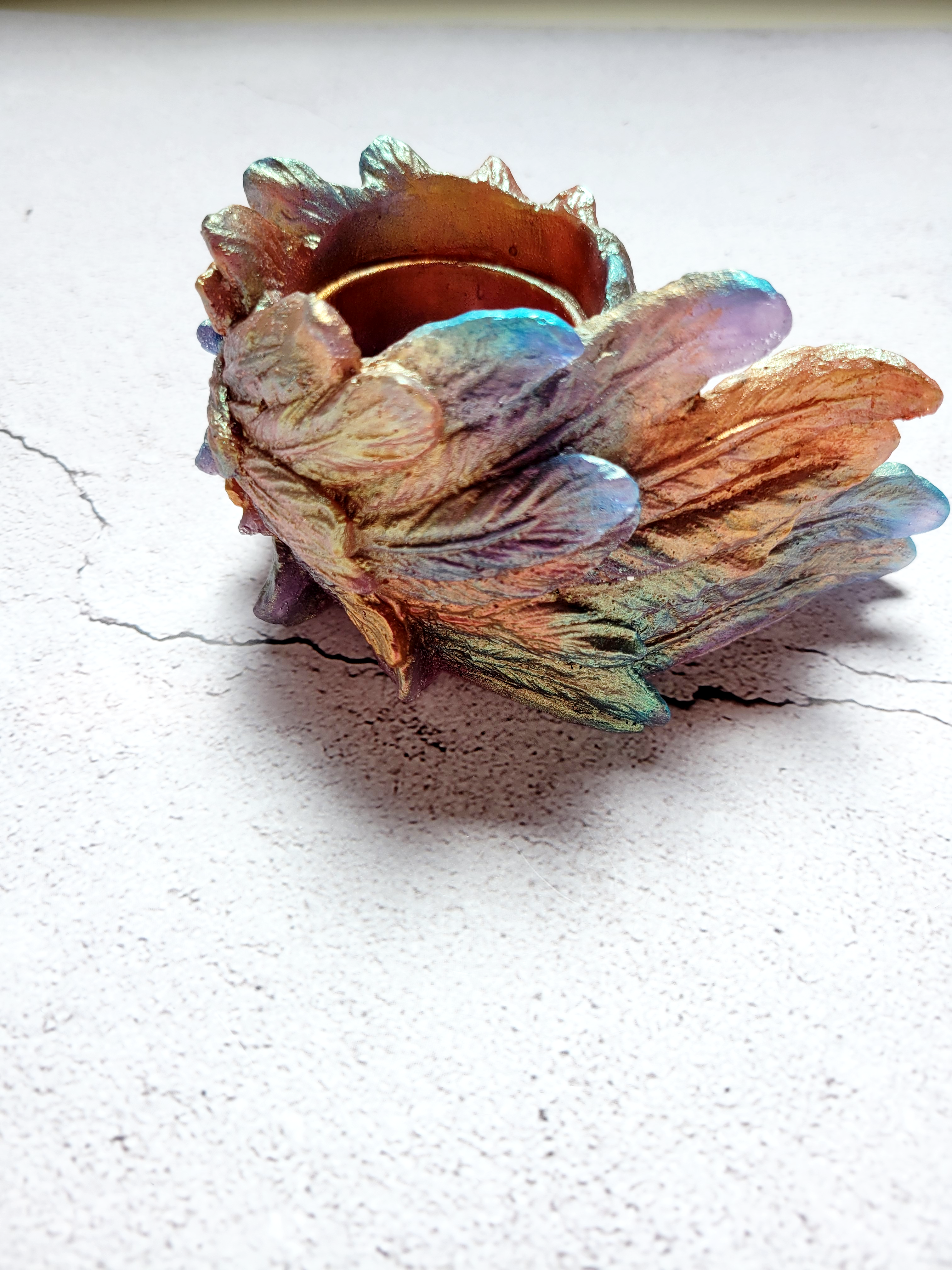 A side view of a tealight candle holder in the style of a bird's wing. It's textured feathers are various colors of blues, oranges, golds, greens.