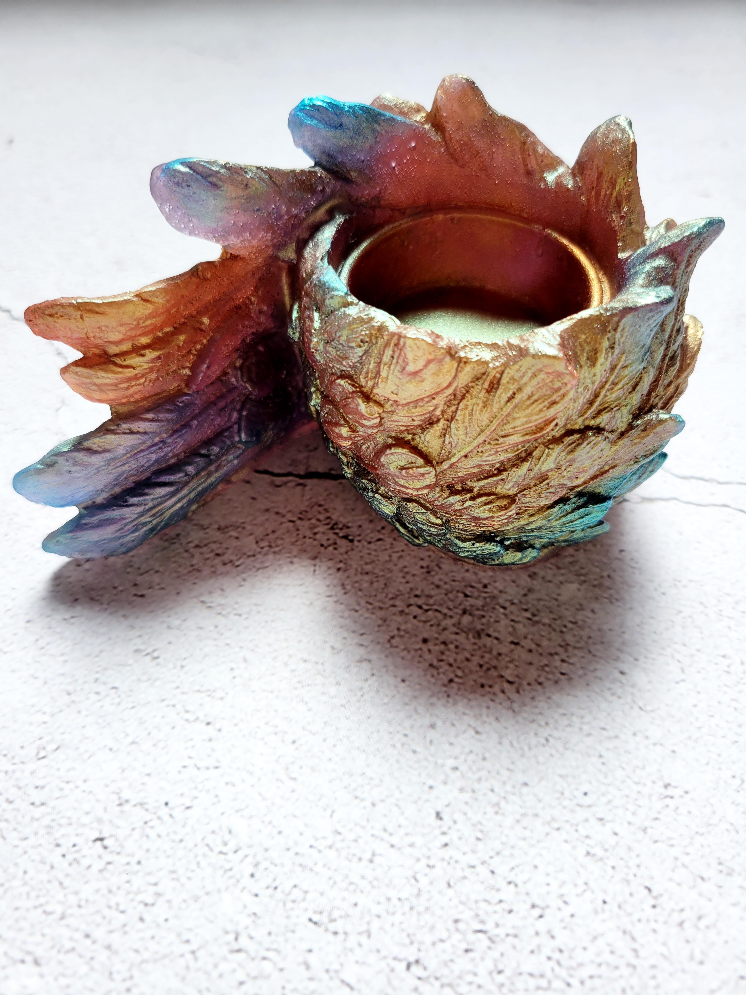 A side view of a tealight candle holder in the style of a bird's wing. It's textured feathers are various colors of blues, oranges, golds, greens.