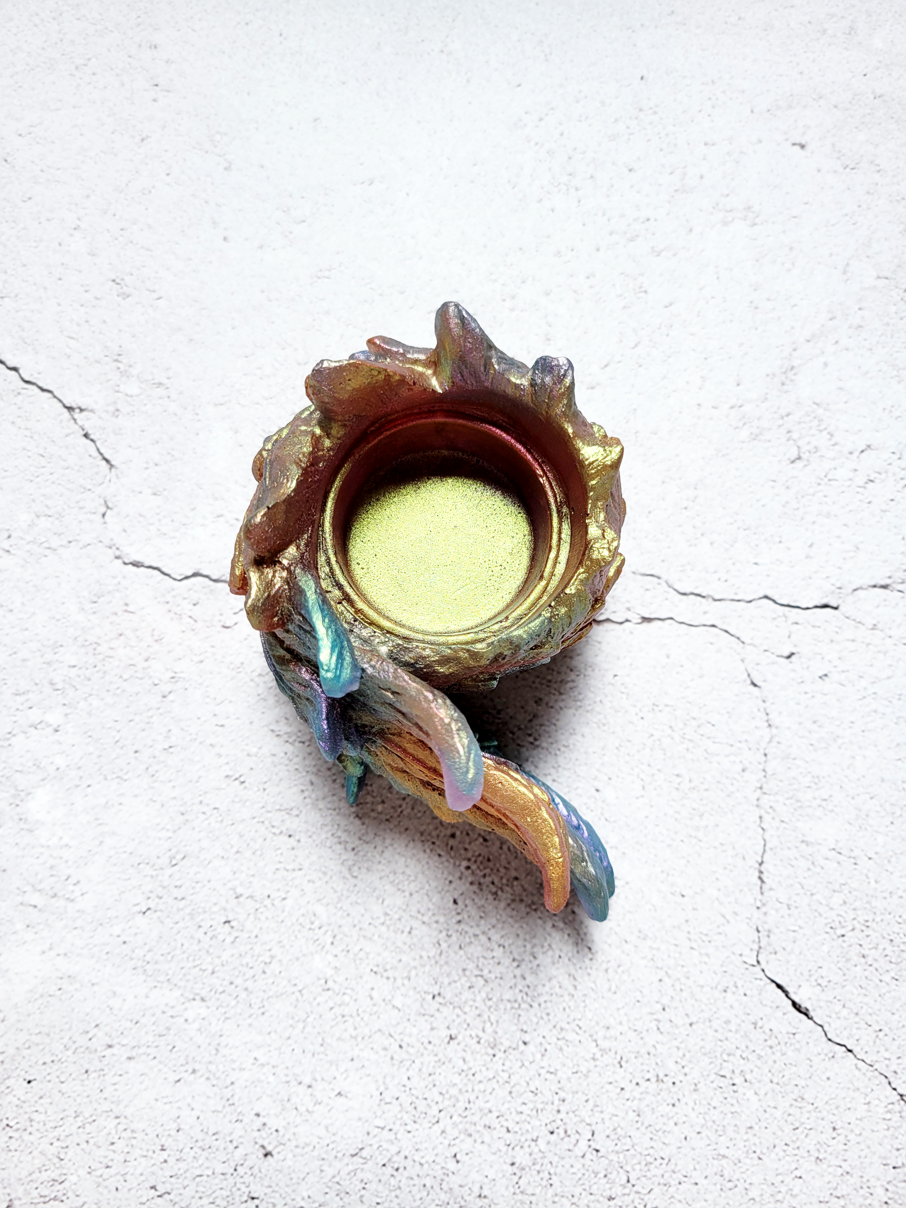 A birds eye view of a tealight candle holder in the style of a bird's wing. It's textured feathers are various colors of blues, oranges, golds, greens.