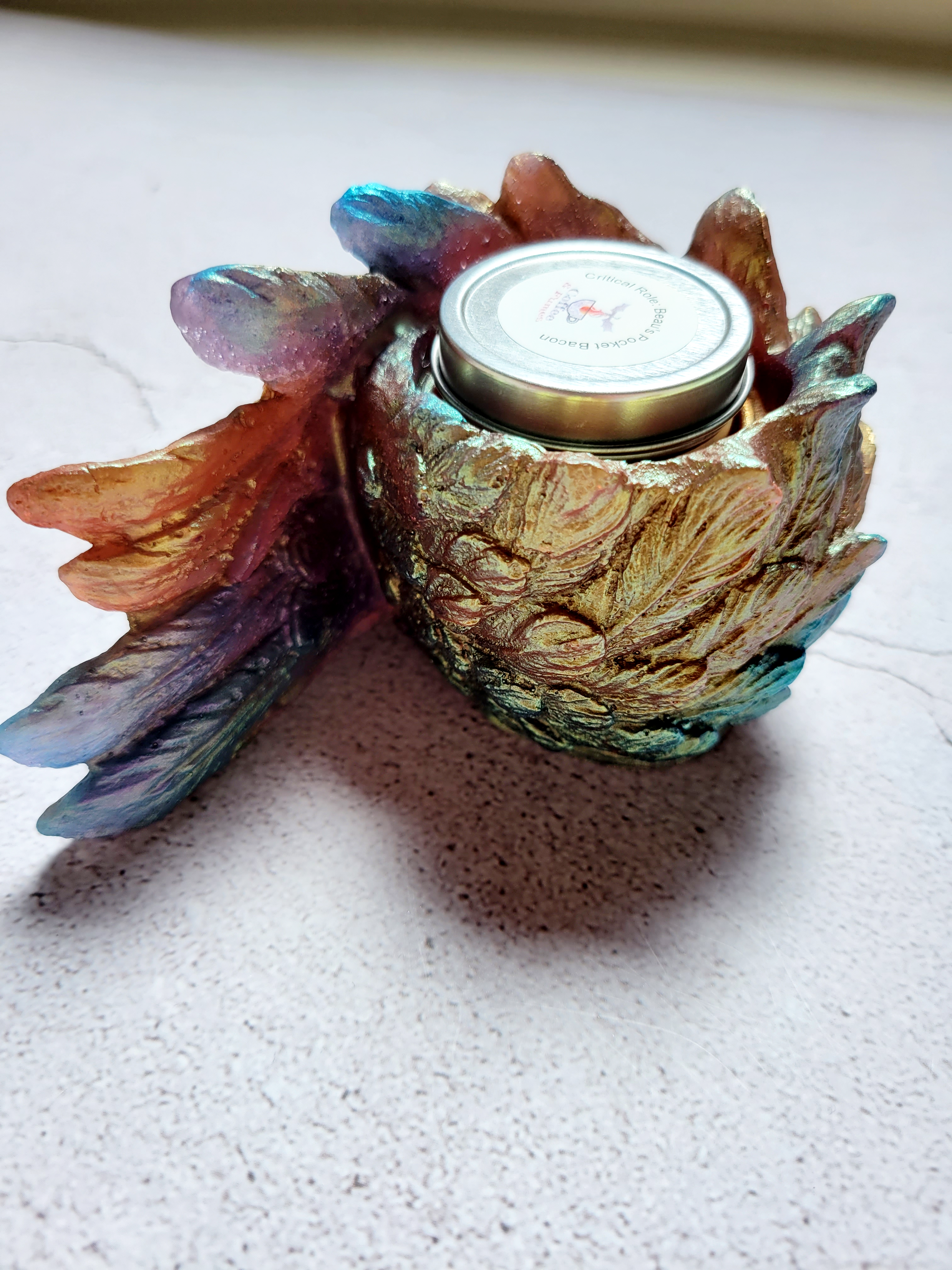 A side view of a tealight candle holder in the style of a bird's wing. It's textured feathers are various colors of blues, oranges, golds, greens. There is a tealight candle inside to show size comparison.