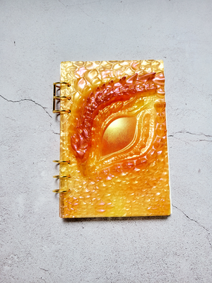 A top view of a closed textured dragon eye journal/notebook. It has two three-ring gold clasps and some A5 paper. The notebook is predominately yellow with gold and red highlights. 