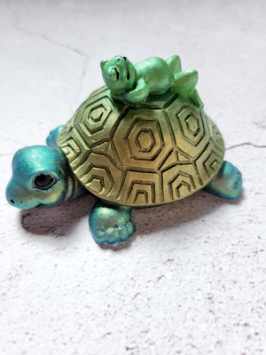 A side view of a turtle and frog figure. The turtle is dark green with green shell, black painted eyes and nostrils. The frog is lounging on the top of the shell. The frog is bright green with black painted features.