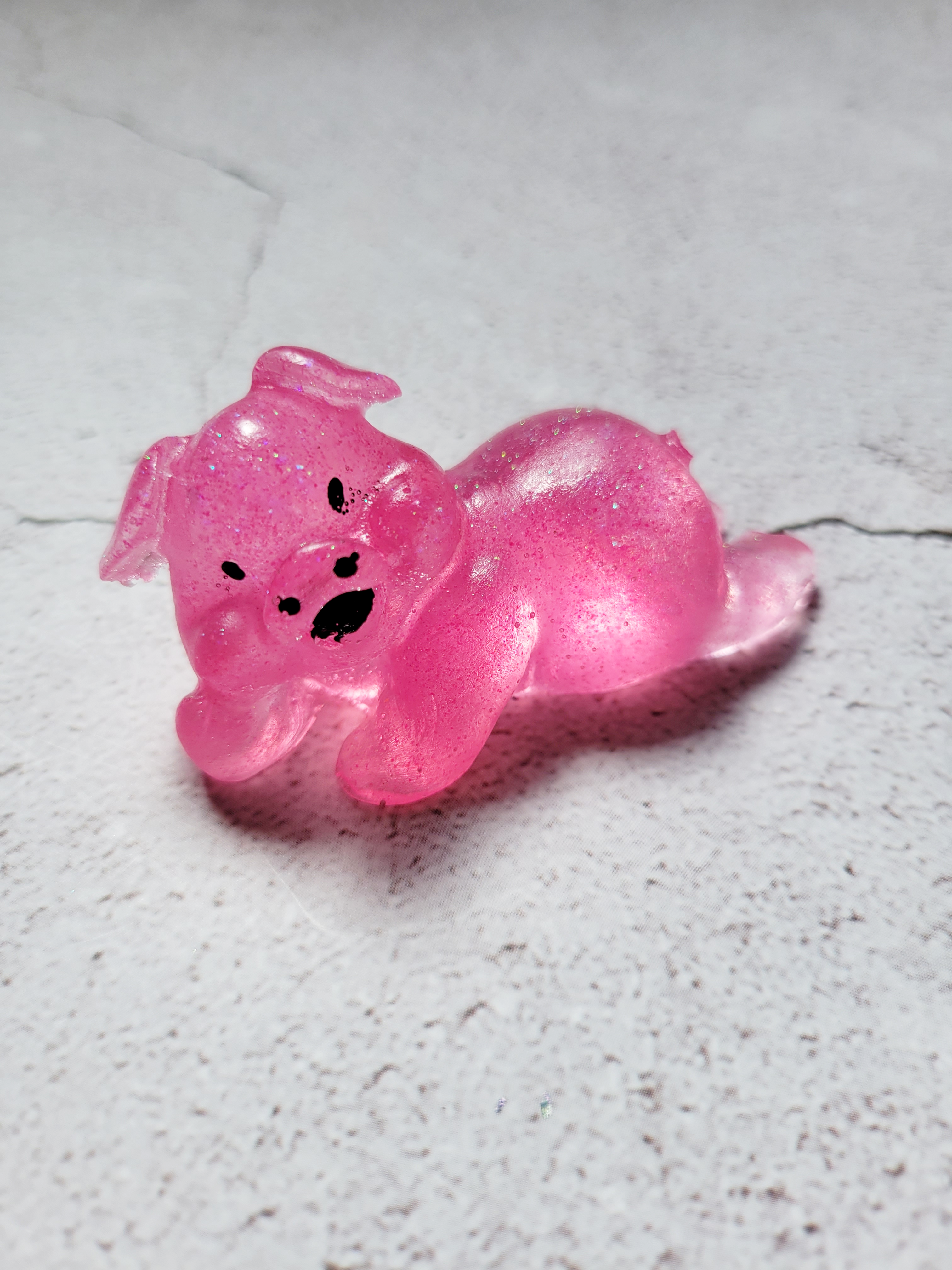 A front view of a lounging pig figure. It has black painted eyes, nose, and mouth. It's pink with glitter throughout.