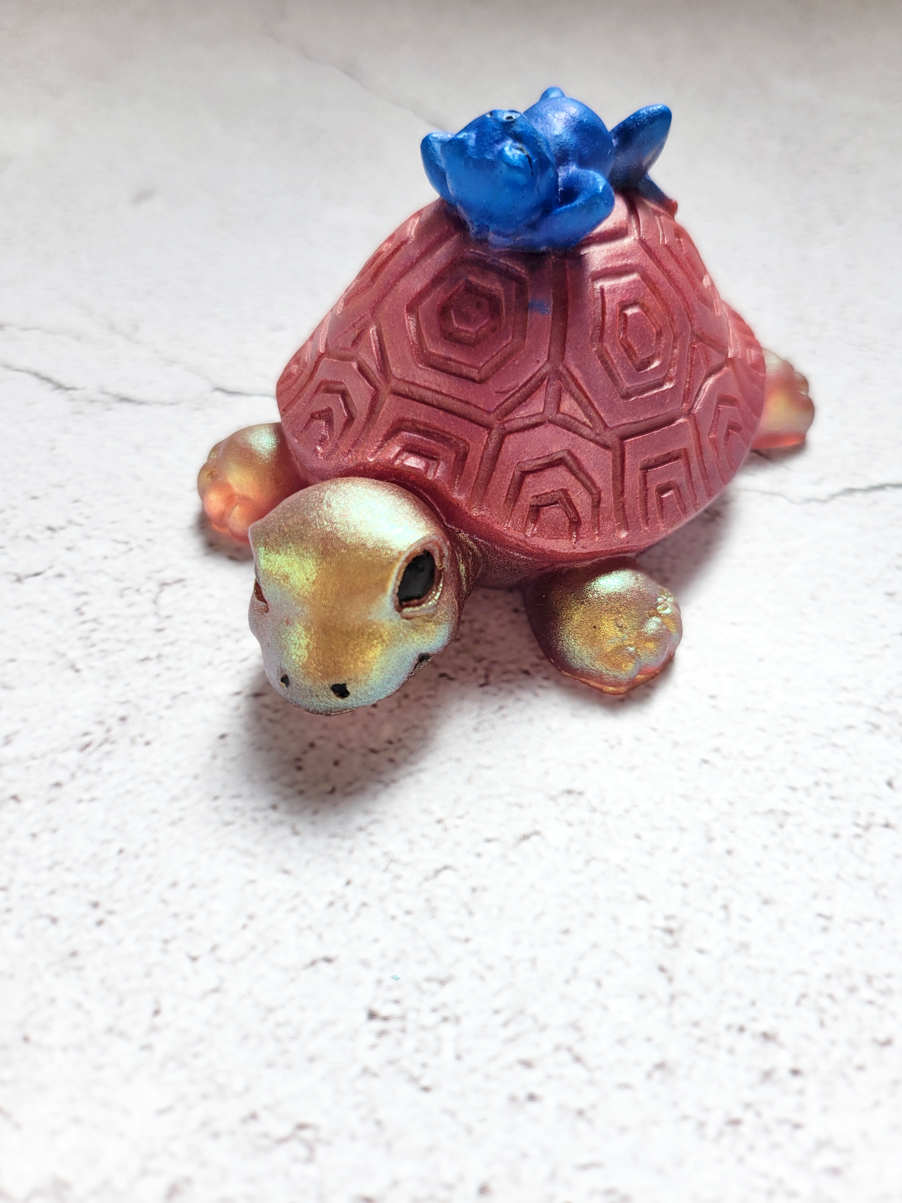 A front view of a turtle figure with a blue frog lounging on top of its shell. The turtle is gold with a reddish shell, with black eyes and nostrils.