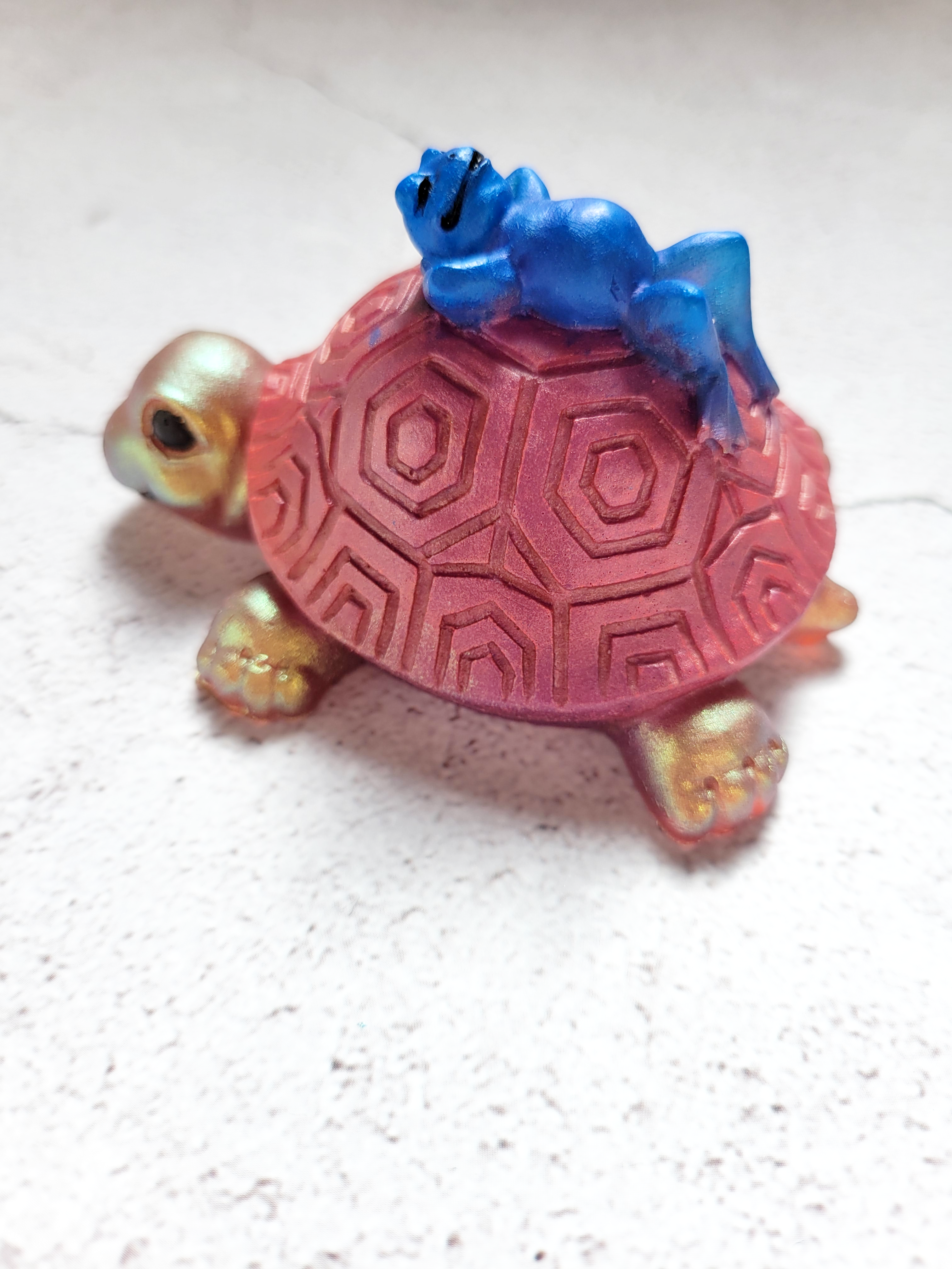 A side view of a turtle figure with a blue frog lounging on top of its shell. The turtle is gold with a reddish shell, with black eyes and nostrils.