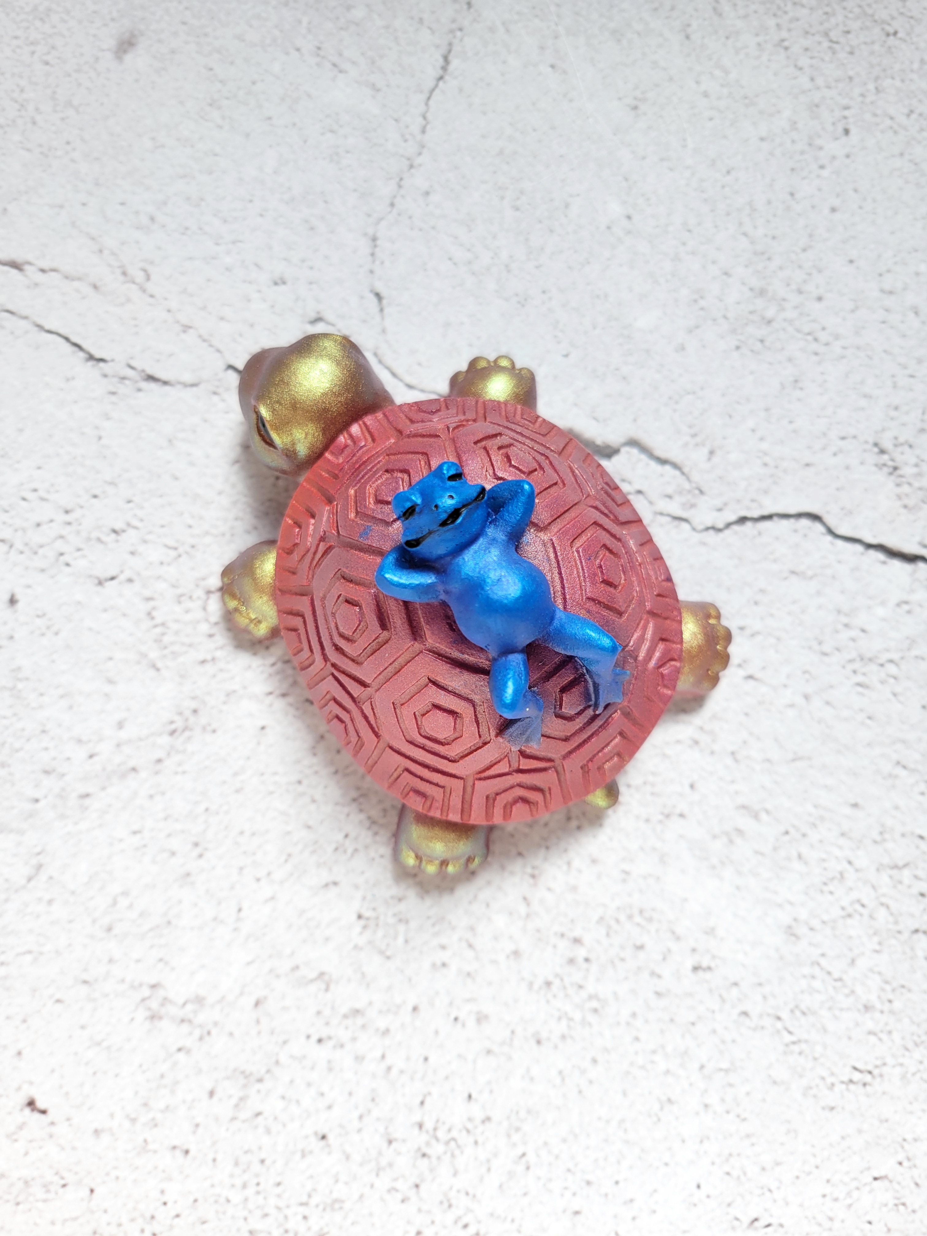A top view of a turtle figure with a blue frog lounging on top of its shell. The turtle is gold with a reddish shell, with black eyes and nostrils.