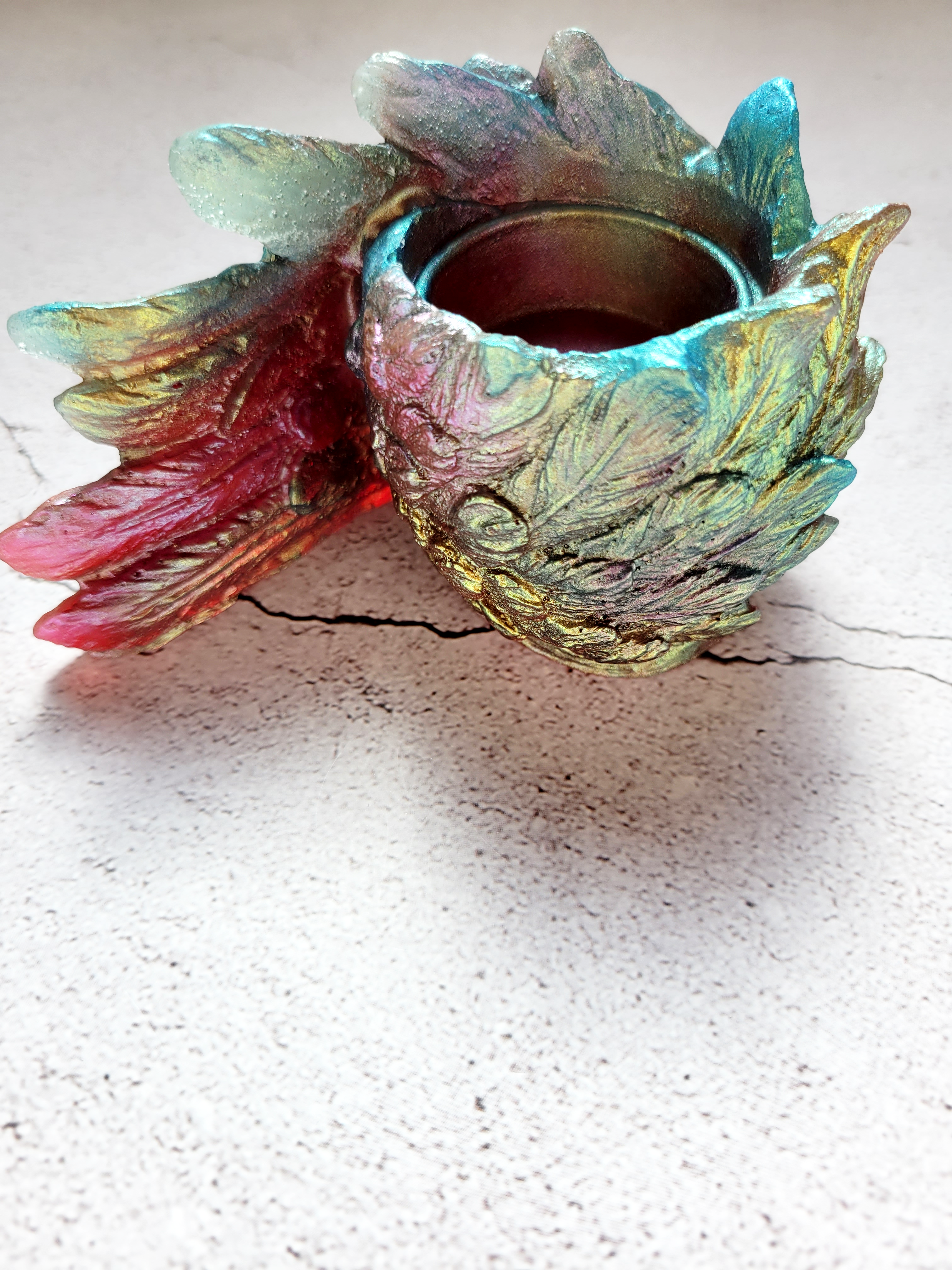 A side view of a tealight candle holder in the style of a bird's wing. It's feathers a textured. It's colorshifting reds, greens, blues.