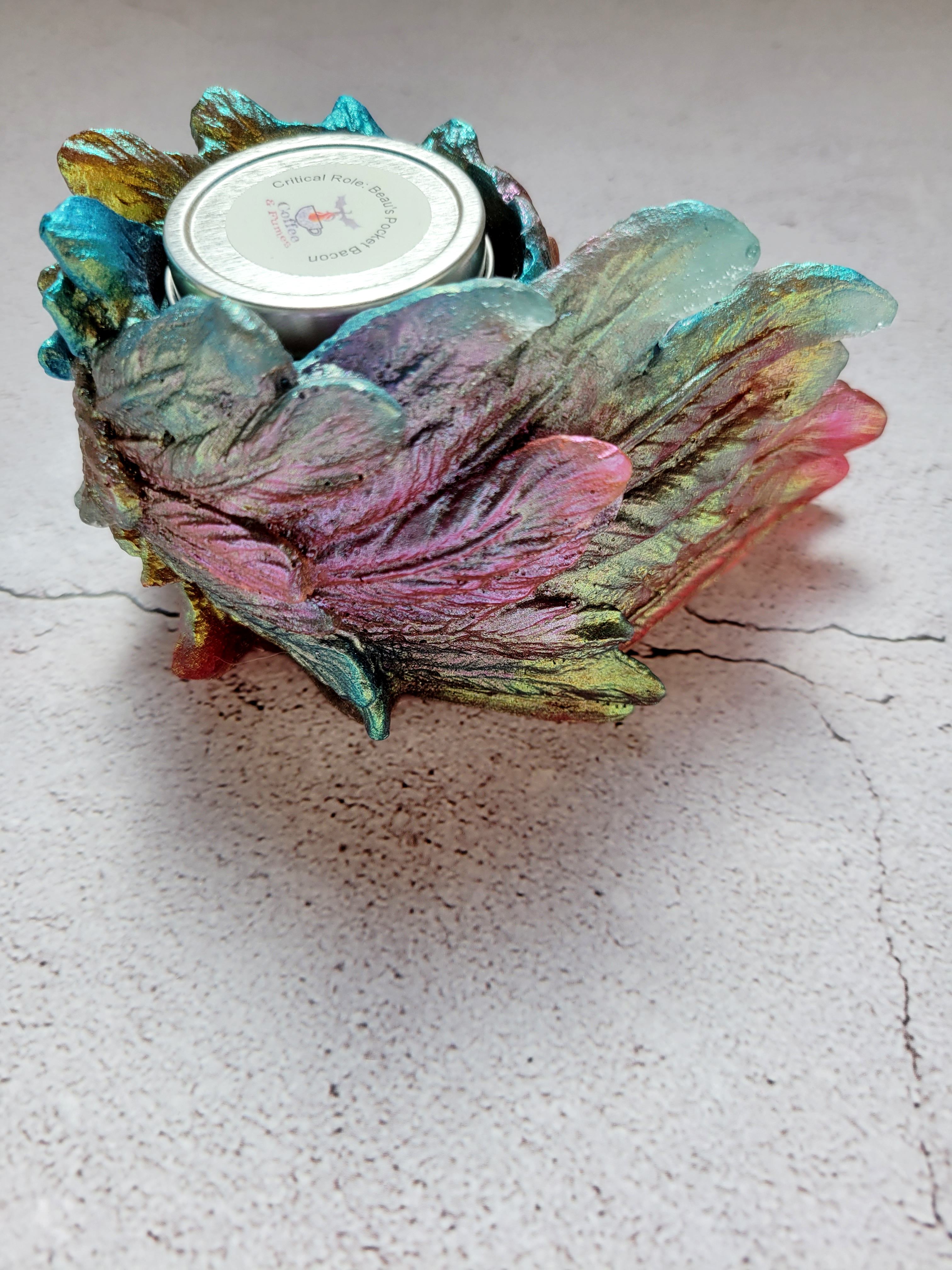 A side view of a tealight candle holder in the style of a bird's wing. It's feathers a textured. It's colorshifting reds, greens, blues. There's a tealight inside to show size comparison.