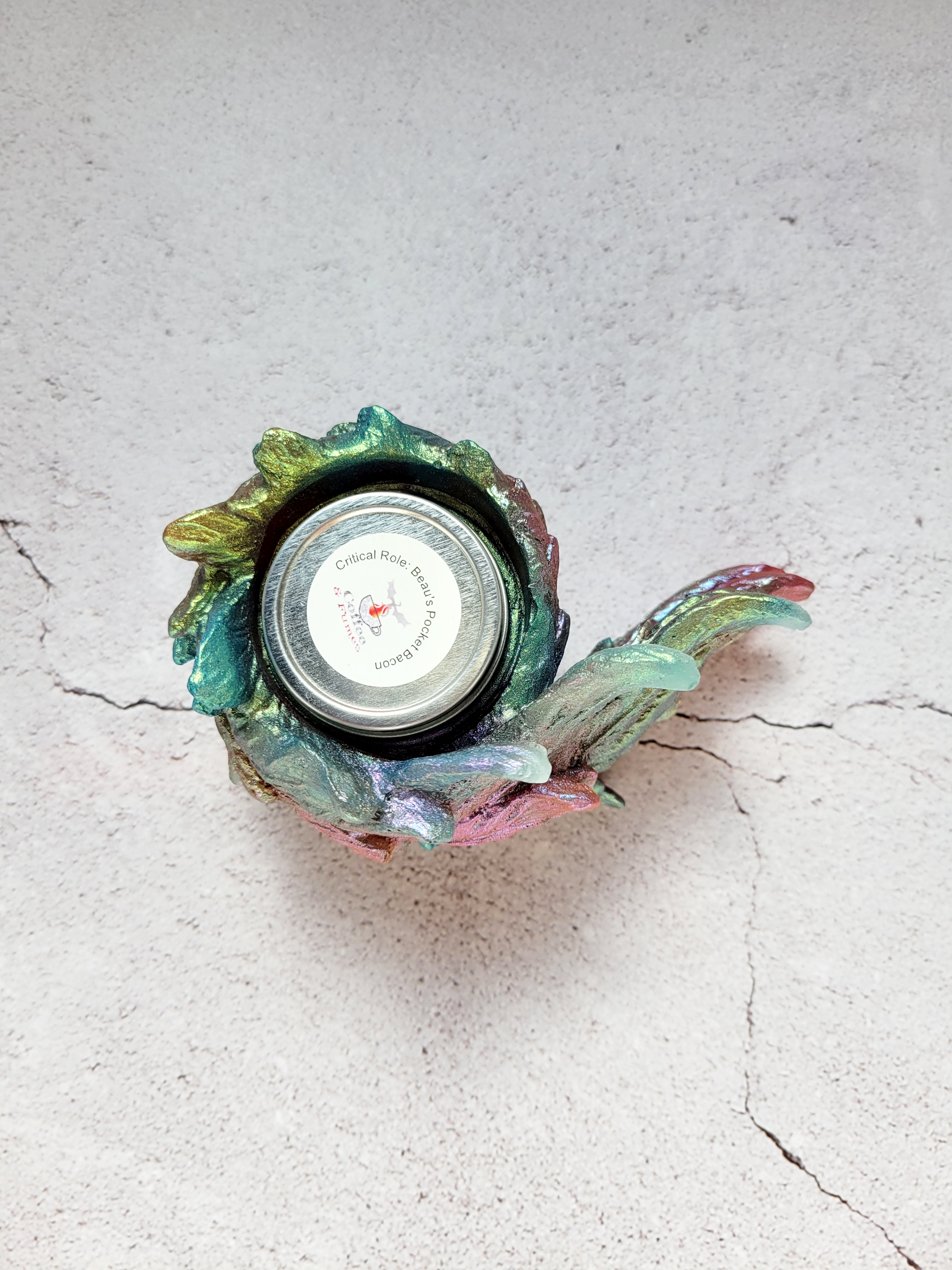 A top view of a tealight candle holder in the style of a bird's wing. It's feathers a textured. It's colorshifting reds, greens, blues. There's a tealight inside to show size comparison.