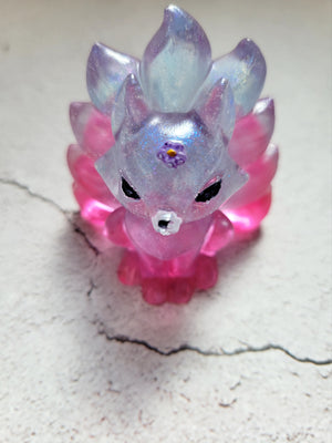 a resin made kitsune fox figure with black painted eyes, purple painted flower on its head. the body is a blend of blue and pink. front view