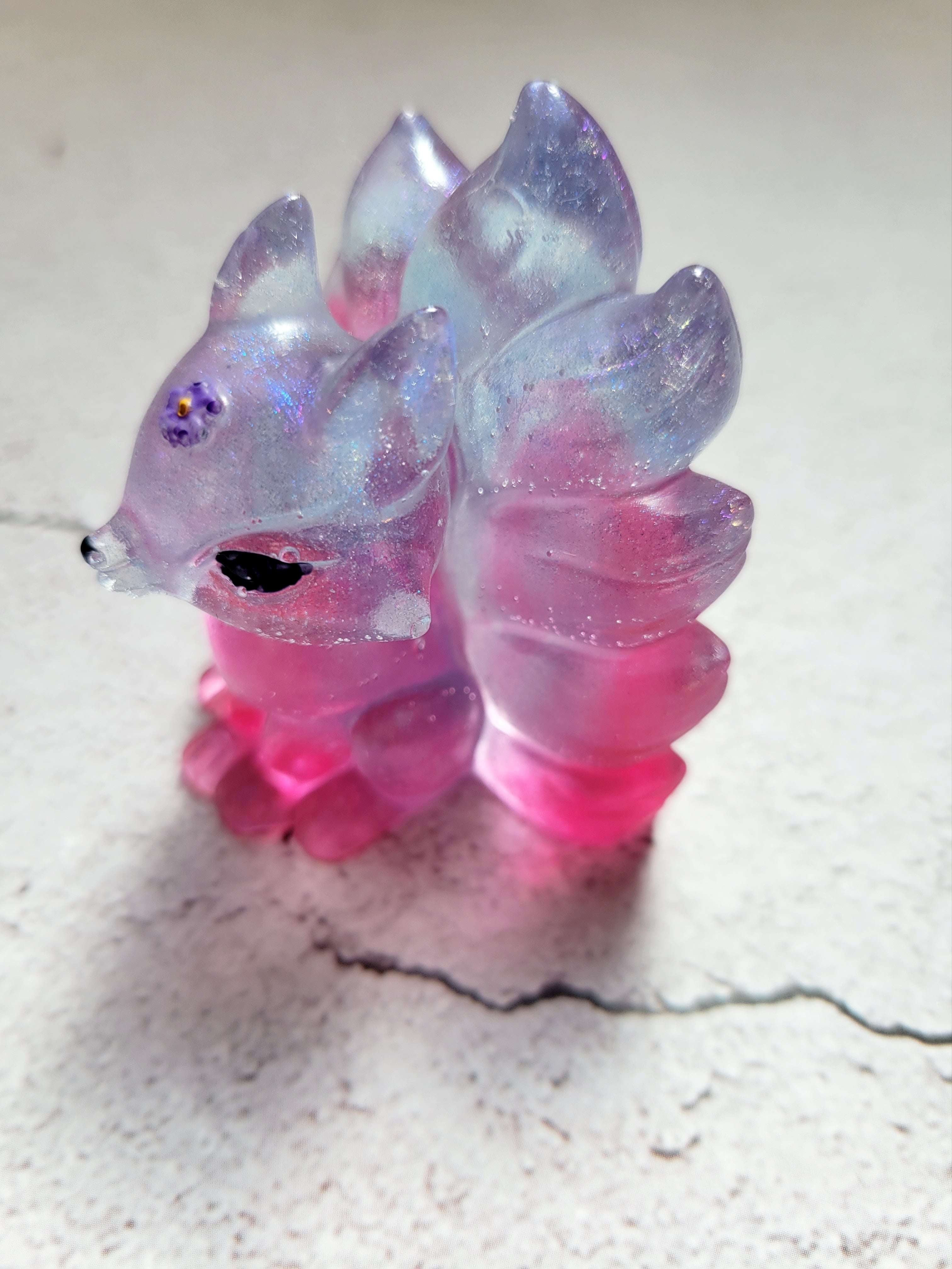 a resin made kitsune fox figure with black painted eyes, purple painted flower on its head. the body is a blend of blue and pink. side view