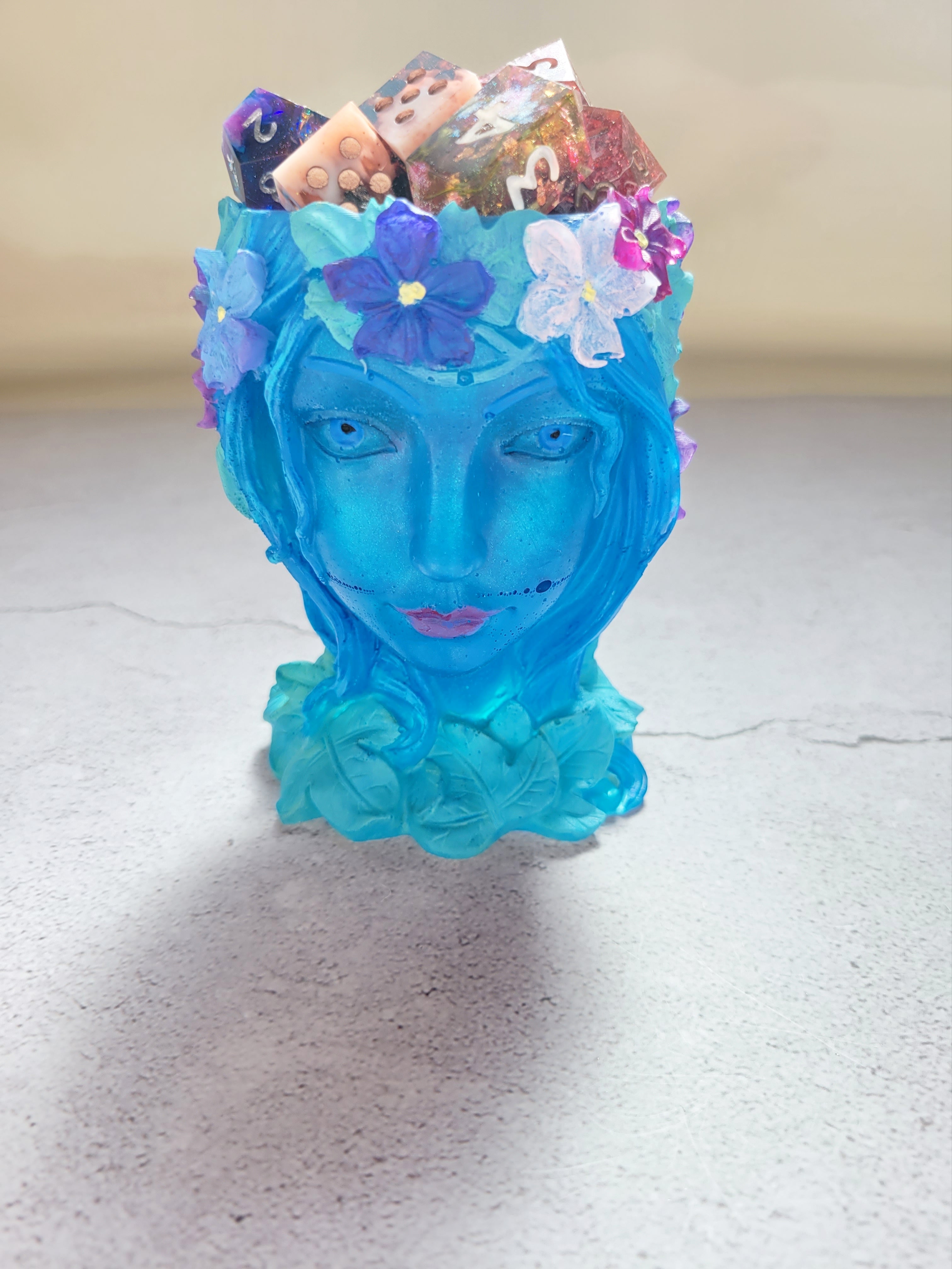 a resin made female ancient statue bust, blue in color with painted leaves and flowers. front view with dice in the holder