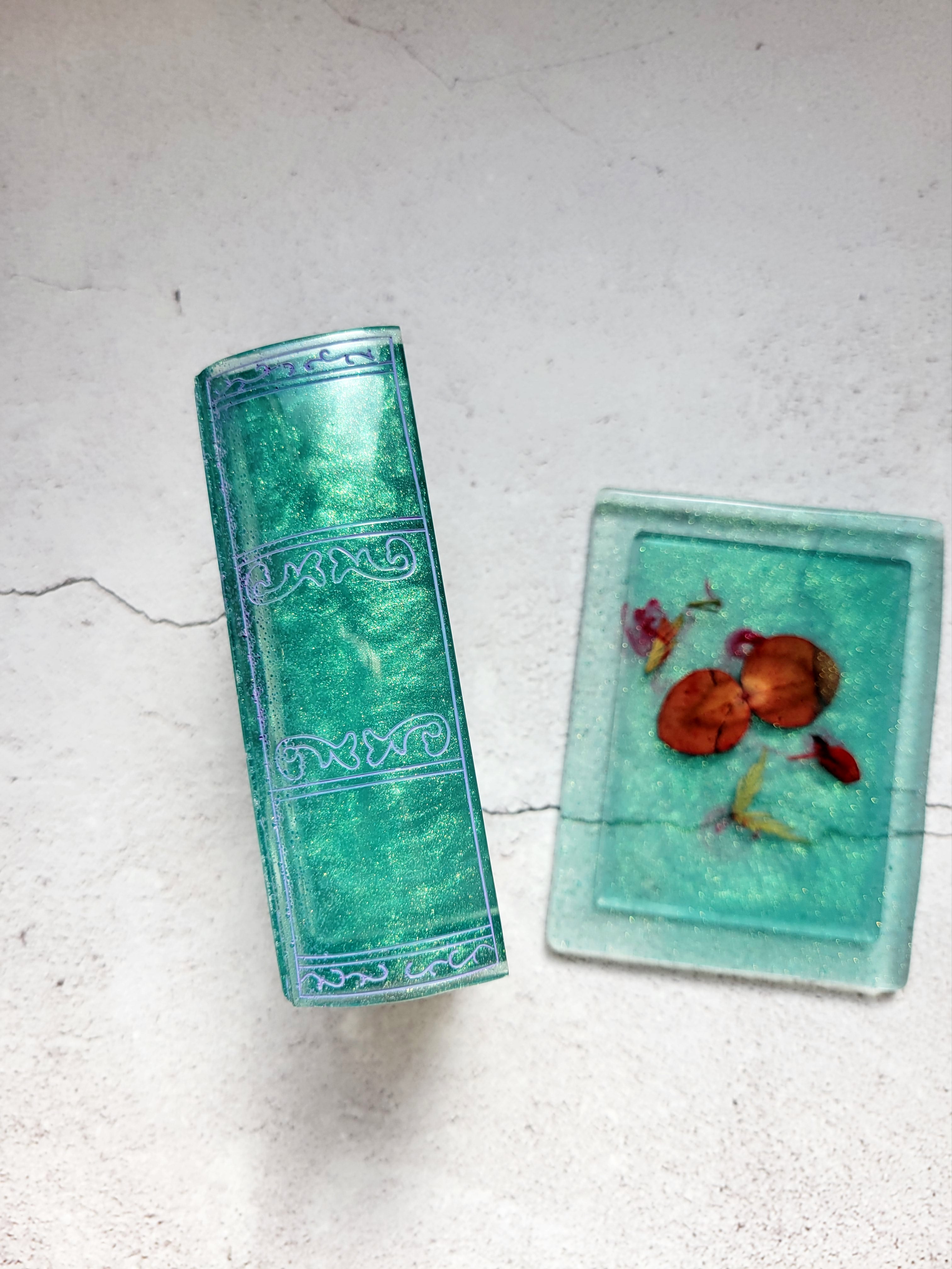a resin made dice vault in the style of a book. The lid is beside the box to show the design of plant leaves. The box is on its side to show the purple painted embellishments on the spine.  It's a shimmering green color.