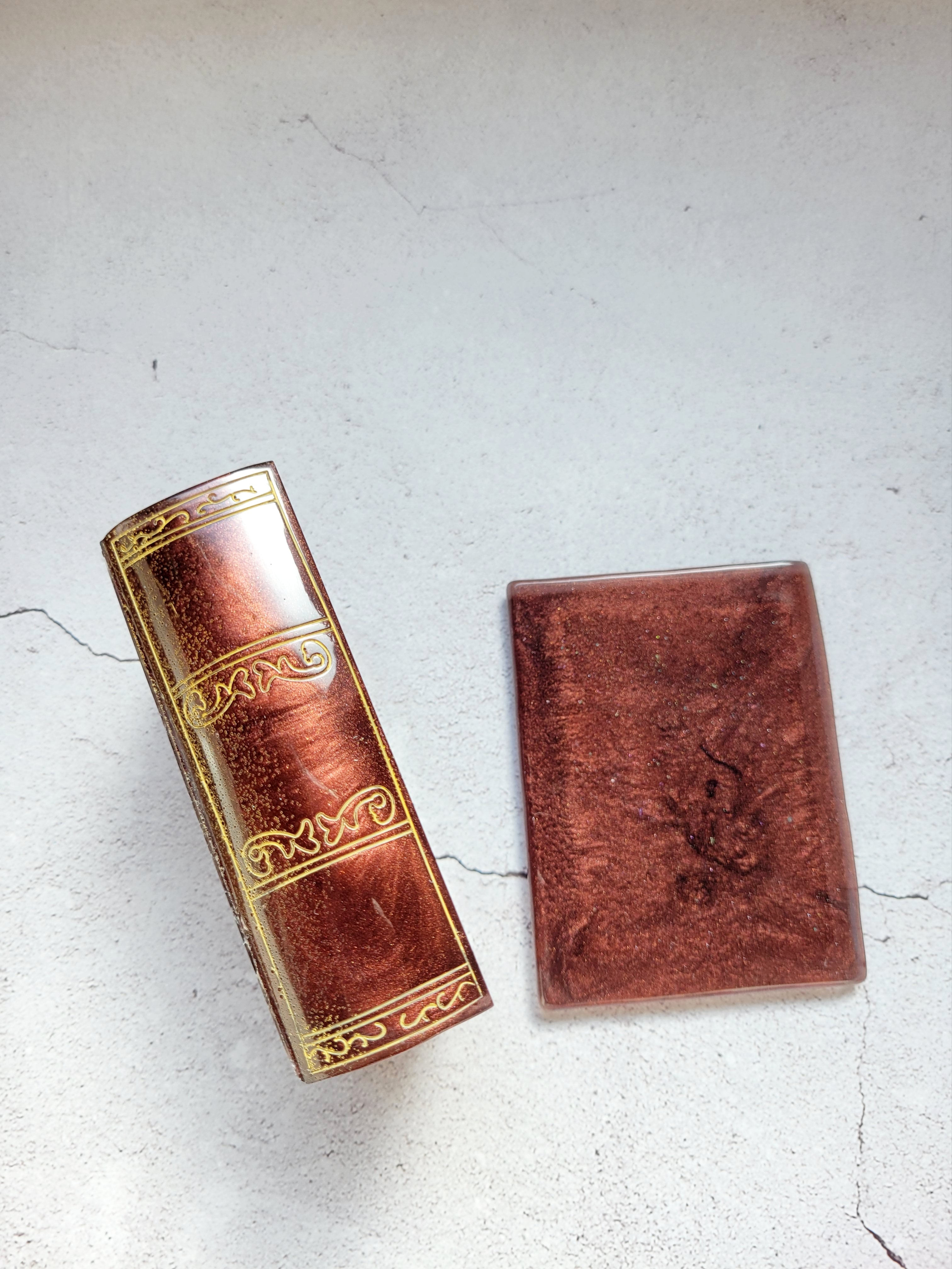 a resin made dice tome box, the lid is beside the box, the box is on its side to show the gold embellishments on the spine. . It's a shimmering brown and black color.