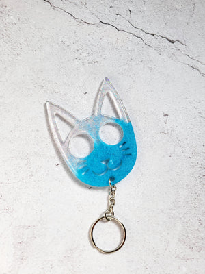 A defense keychain in the style of a cat face. It has a silver chain and ring hoop. It's clear and blue with glitter.