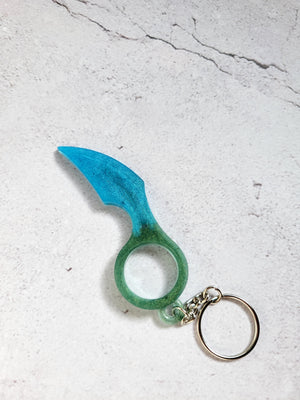 A defense keychain with a silver chain and ring hoop. It has a curved edge. It's blue and green.